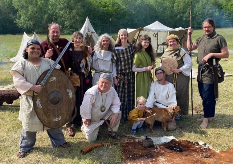 Gruppe von Menschen in historischer Kleidung vor mittelalterlichem Lager mit Zelten. Einige tragen Waffen oder Schilde, ein Kind und ein Hund sind ebenfalls zu sehen.