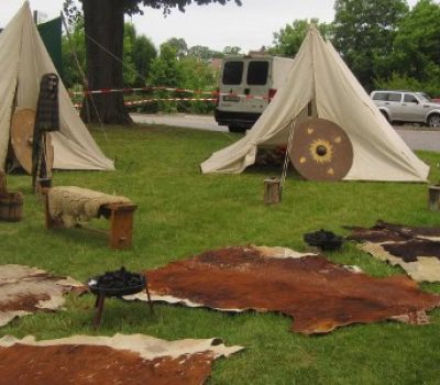 Aufbau eines kleinen Germanenlagers mit weißen Zelten, bemalten Rundschilden, Fellen auf dem Gras und einfachen Feuerstellen – Darstellung historischer Lebensweise durch den Verein Sugambrer e.V. im Freien.